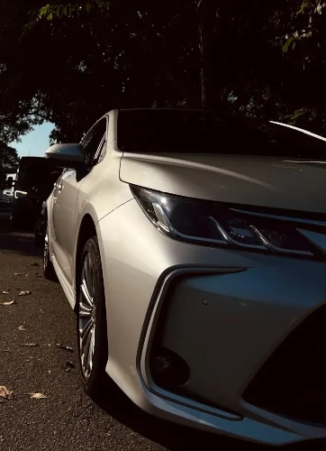 Carro sedan Corola prateado estacionado à sombra de árvores em rua asfaltada